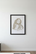 A white art print featuring a sepia-toned sketch portrait of Bob Marley with the text 'BOB MARLEY' and a small leaf emblem below, photographed resting on a gray textured fabric surface.