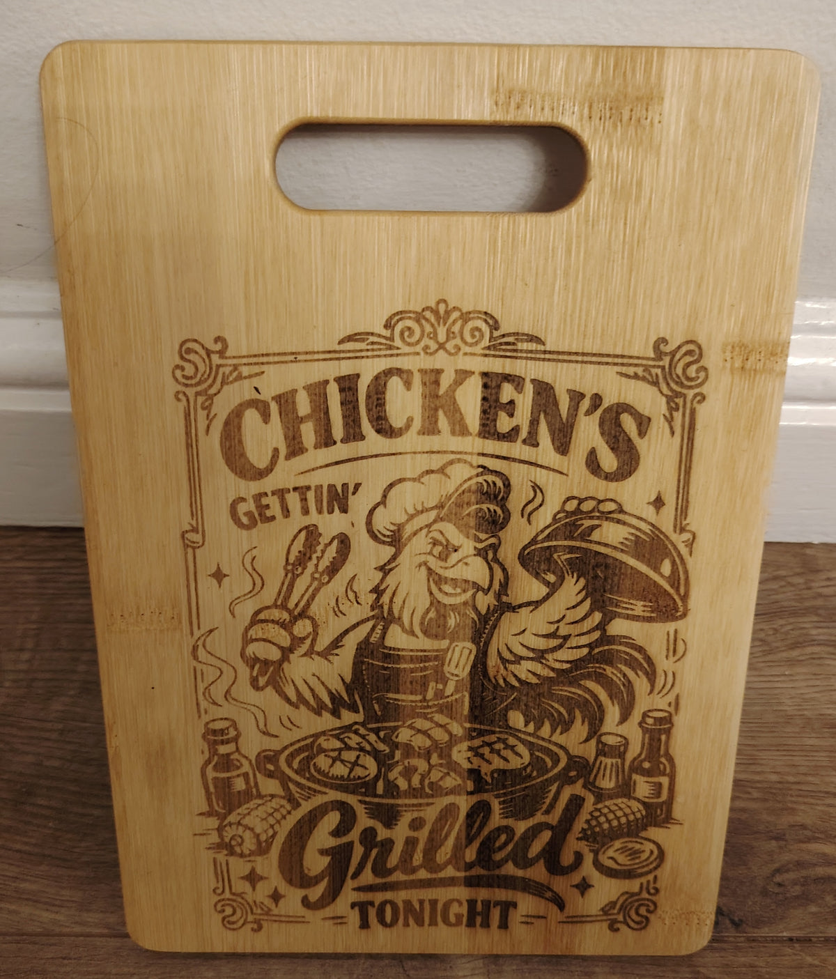 Wooden cutting board with a engraved design of a chef and chicken, text 'Chicken's Gettin' Grilled Tonight', on a wooden surface.