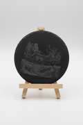 A round black slate coaster featuring an embossed design of Santa Claus in a sleigh pulled by reindeer over a mountain landscape, displayed on a small wooden easel against a light gray background.