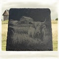 A rectangular dark slate plaque featuring a detailed engraved or printed depiction of a Highland cow in a field with a barn in the background, displayed outdoors on a grassy field with a cloudy sky.