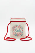 A small cream canvas crossbody bag with a red zipper and red strap, featuring a floral wreath design and the name 'Enola' embroidered in teal on the front, set against a gray textured background.
