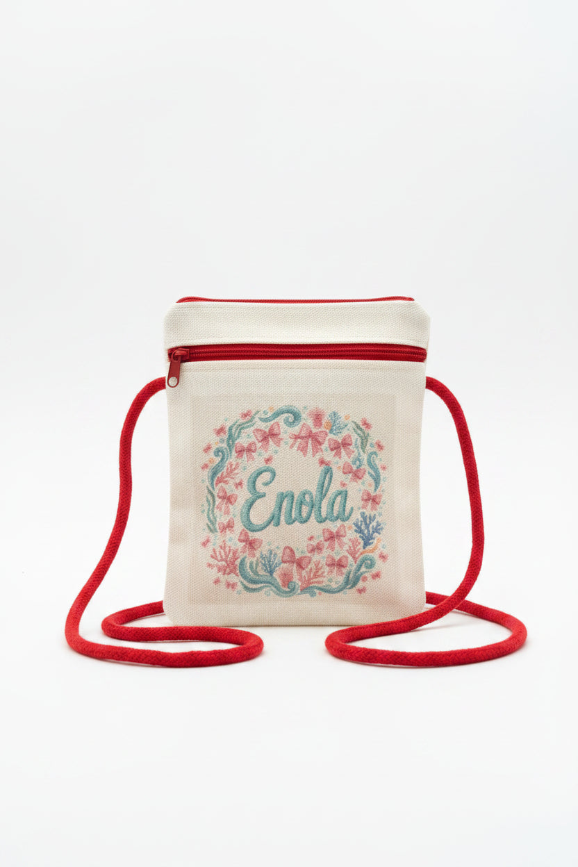A small cream canvas crossbody bag with a red zipper and red strap, featuring a floral wreath design and the name 'Enola' embroidered in teal on the front, set against a gray textured background.