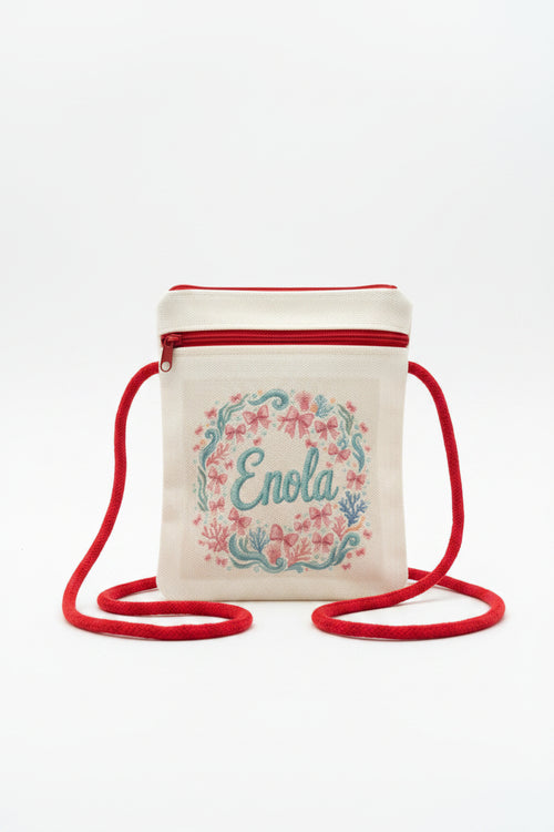 A small cream canvas crossbody bag with a red zipper and red strap, featuring a floral wreath design and the name 'Enola' embroidered in teal on the front, set against a gray textured background.