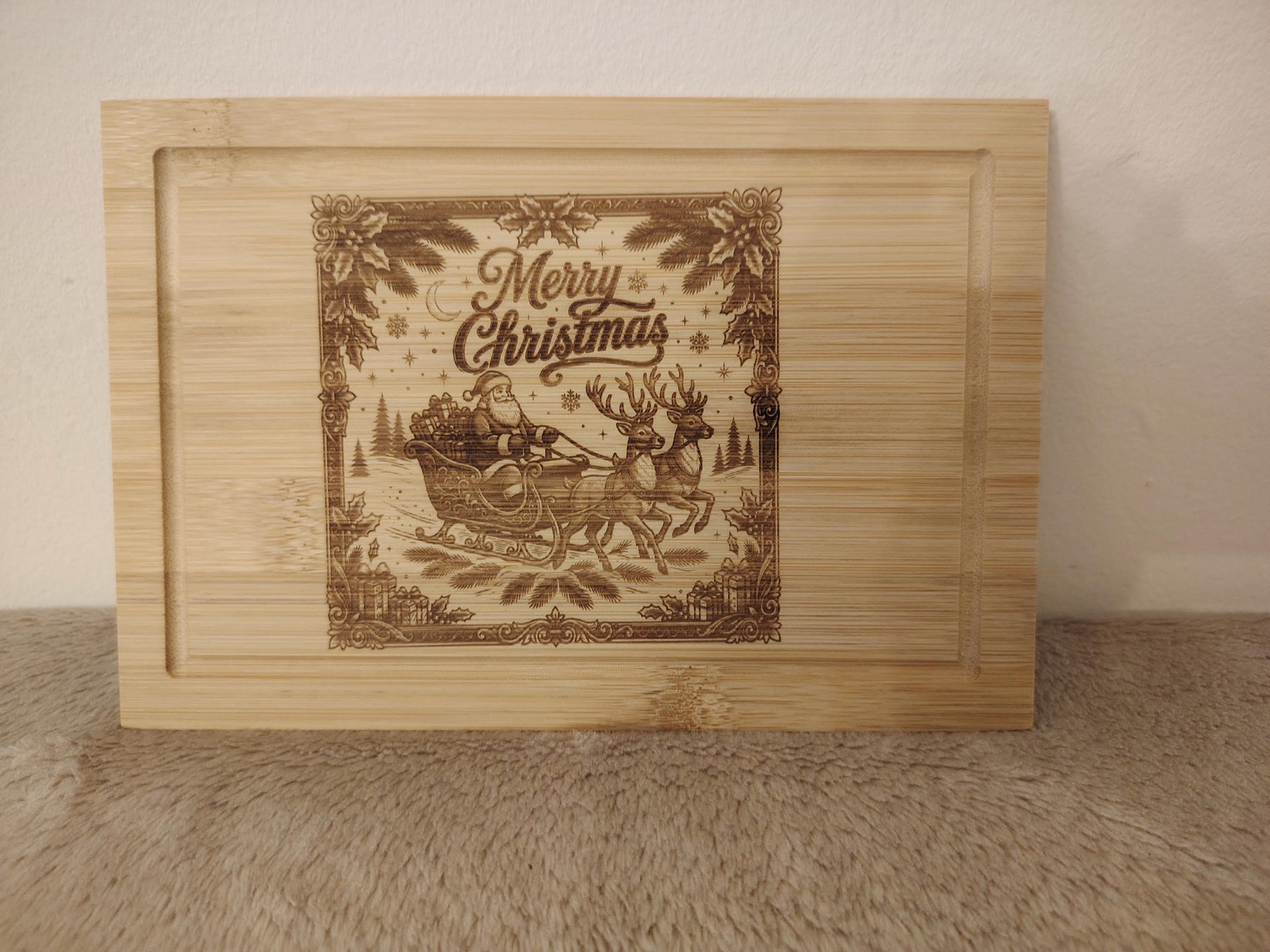 A rectangular light wood tray or cutting board with a recessed border and a laser-engraved Christmas illustration in the center. The design features Santa Claus in a sleigh pulled by reindeer under the words 'Merry Christmas,' surrounded by pine branches and snowflakes.