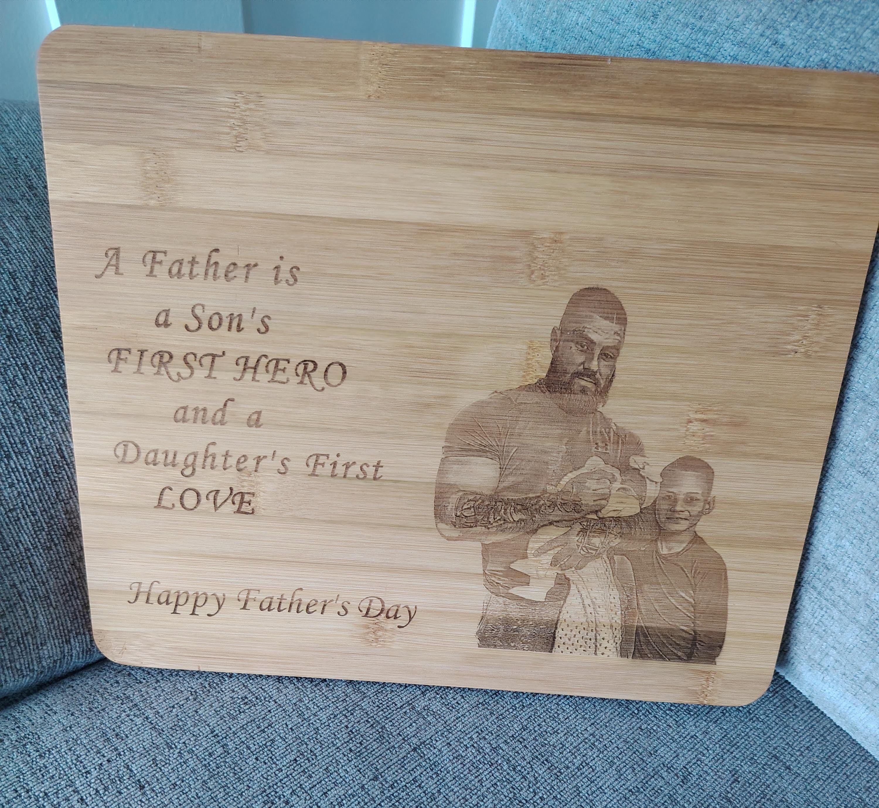 A rectangular wooden Father's Day plaque engraved with the message 'A Father is a Son's FIRST HERO and a Daughter's First LOVE Happy Father's Day' alongside a custom photo of a father and son, resting on a blue textured fabric surface.