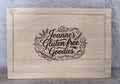 A rectangular light natural wood cutting board with a recessed border and a laser-engraved design that reads 'Joanne's Gluten free Goodies.' in decorative script, shown against a gray textured background.