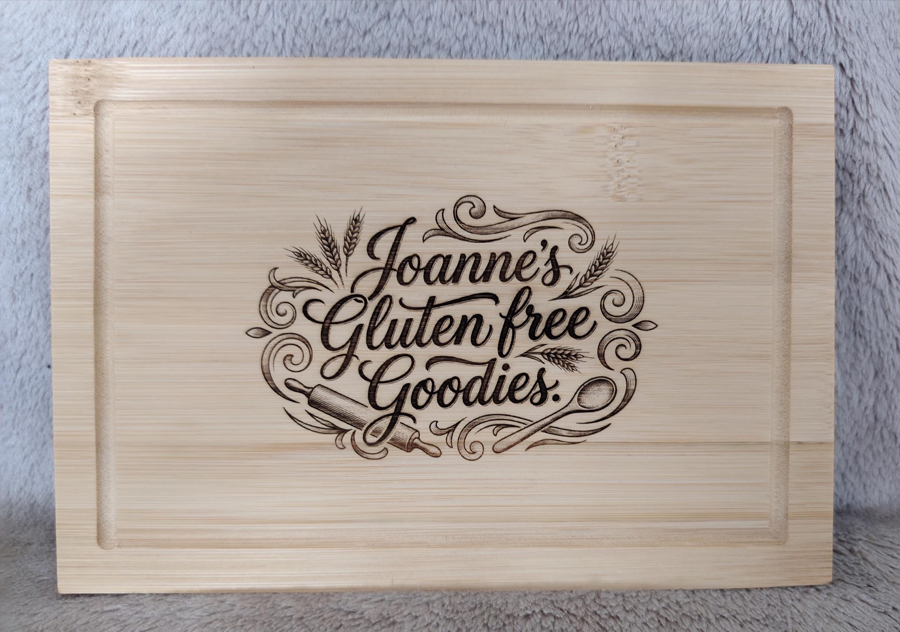 A rectangular light natural wood cutting board with a recessed border and a laser-engraved design that reads 'Joanne's Gluten free Goodies.' in decorative script, shown against a gray textured background.