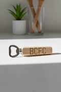 A wooden bottle opener with the letters 'BCFC' engraved on the handle, resting on a textured beige fabric background.