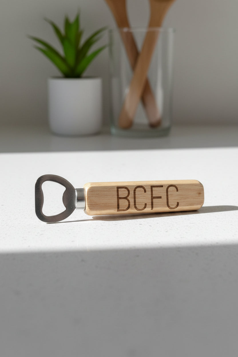 A wooden bottle opener with the letters 'BCFC' engraved on the handle, resting on a textured beige fabric background.