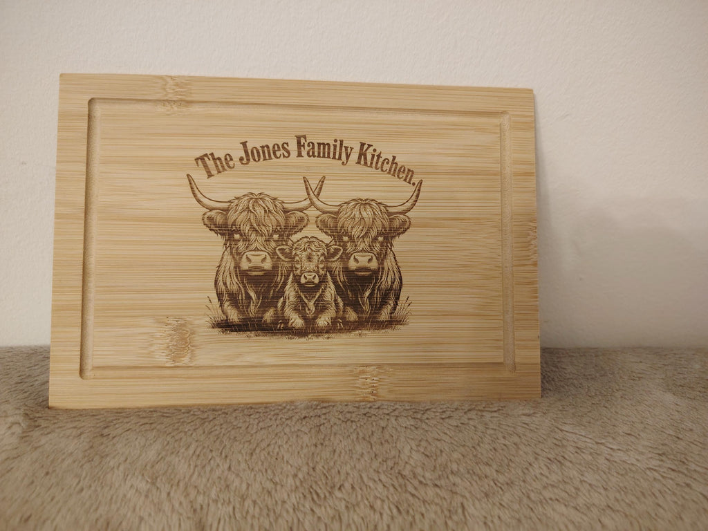A rectangular wooden cutting board with a recessed border, featuring a laser-engraved illustration of three Highland cows and the text 'The Jones Family Kitchen' on a light wood surface, set against a neutral beige background.