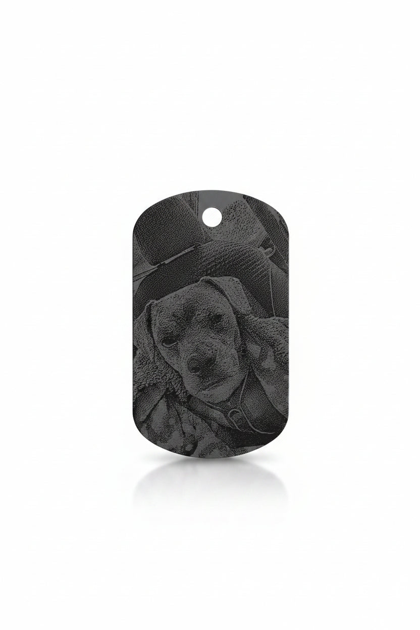 A dog-shaped metal tag with a black and gray engraved portrait of a puppy, resting on a textured beige fabric background.