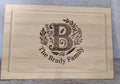 A rectangular light wood cutting board with a recessed center, featuring a custom laser-engraved monogram 'B' with floral details and the text 'The Brady Family' on a gray textured background.