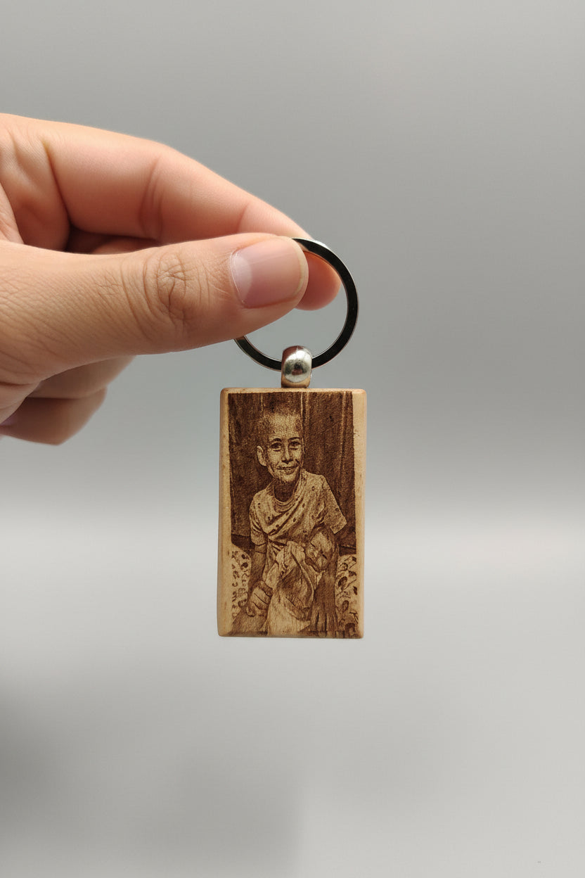 A hand holding a rectangular engraved wooden pendant keychain with a metal split ring. The pendant features a detailed portrait engraving of a smiling person, set against a dark navy blue fabric background.