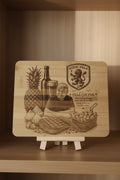 A rectangular wooden board with rounded corners, featuring a laser-engraved design of a pineapple, a bottle, a glass, a lion crest, and a fan Phil graphic, placed on a gray textured fabric surface.