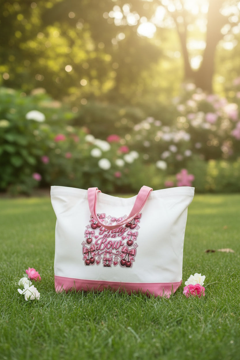 A white canvas tote bag with pink handles and a pink bottom band, featuring a central graphic that reads 'Pretty as a Bow' in pink script surrounded by bows, cherries, and hearts.